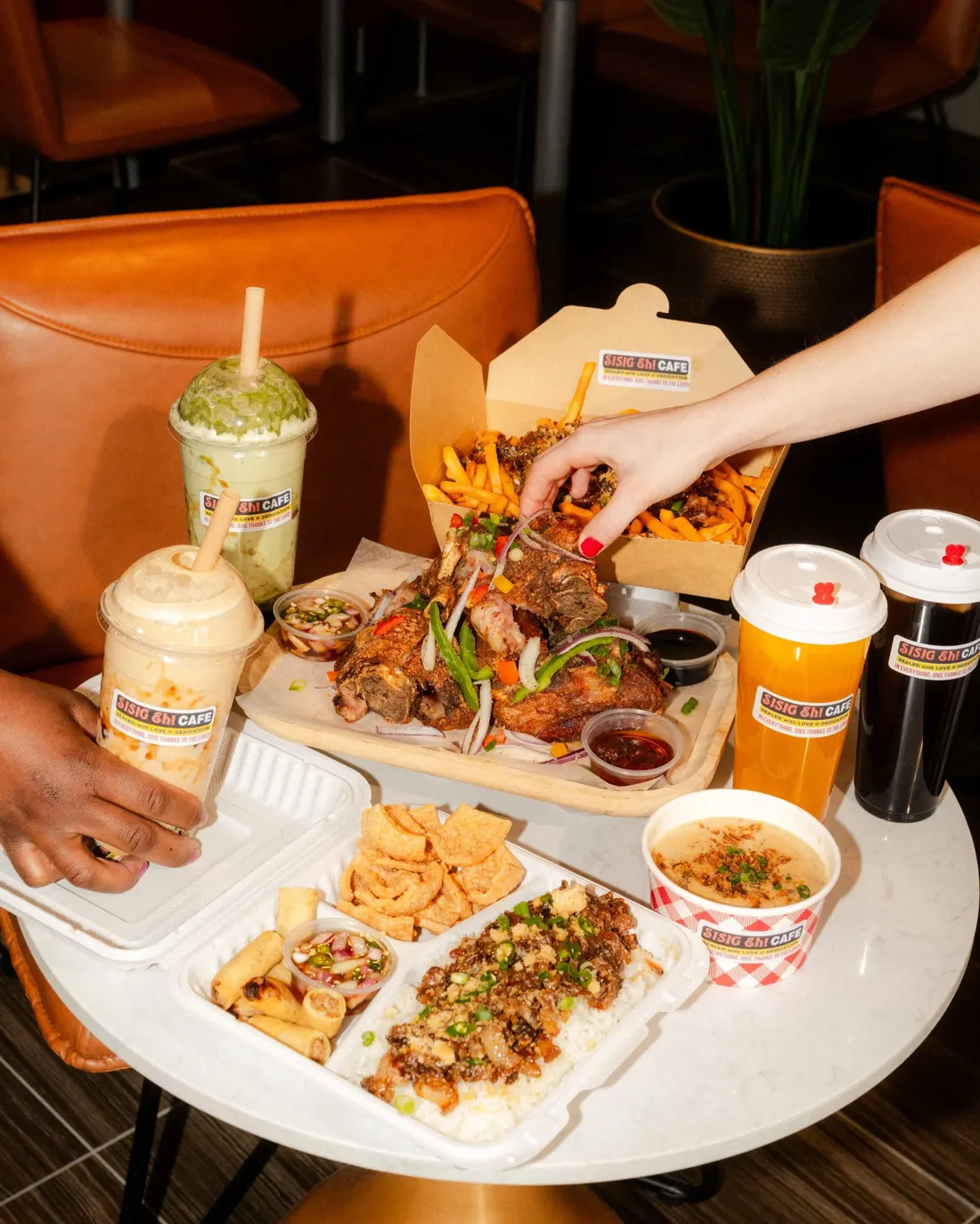 Hands reaching over a tray of grilled meats, sides and iced drinks in takeout containers