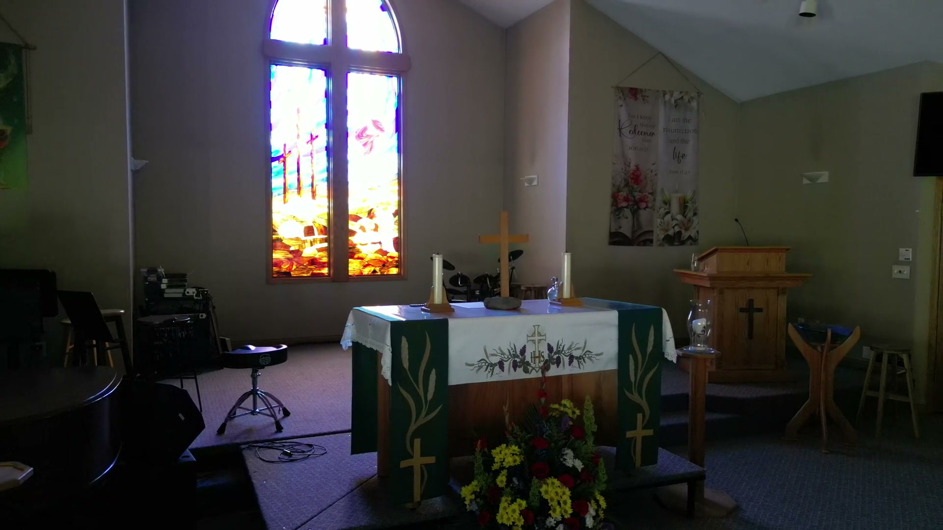 The communion table at St. Peter's drawn back from at half speed: a green frontal worked with wheat and a cross, two lit candles, cut flowers on the step below
