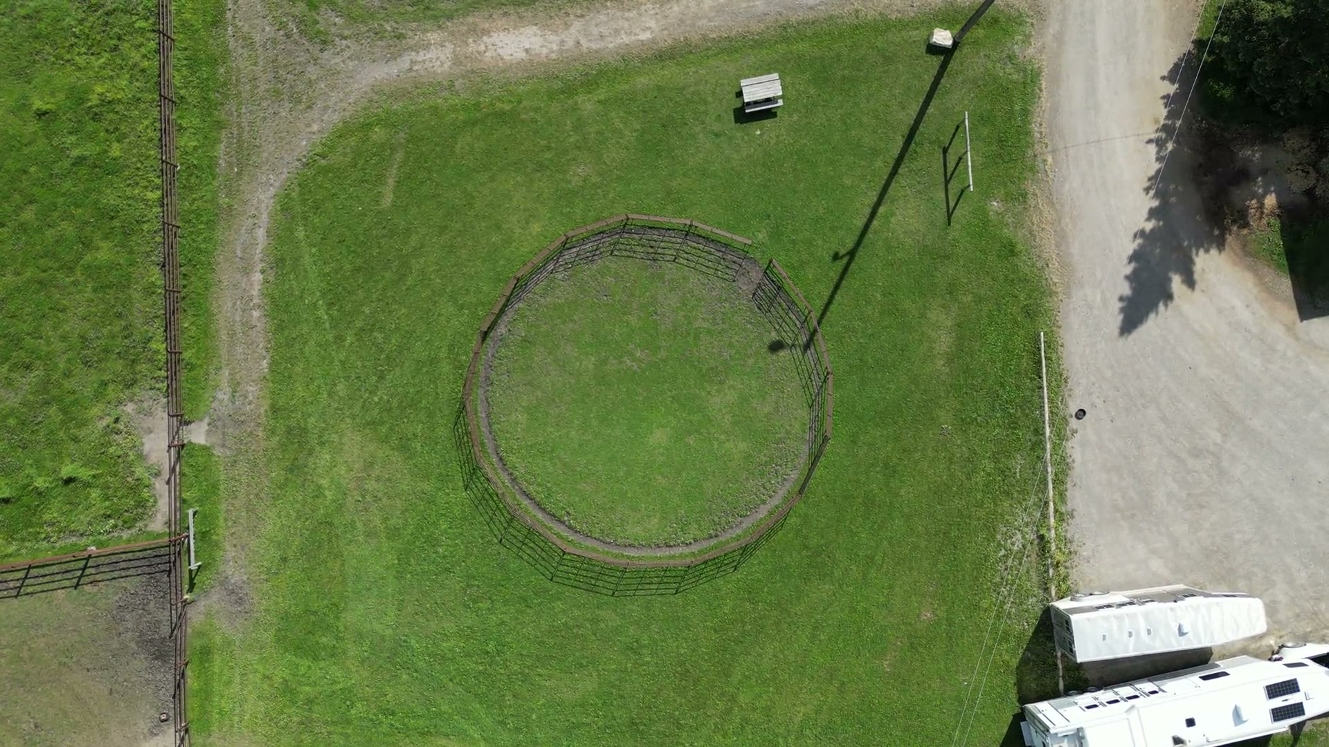 Straight down over a round pen on green grass at Shire Ridge Ranch near Bragg Creek, a gravel lane and parked horse trailers at the edge of the frame, a pole's shadow across the lawn