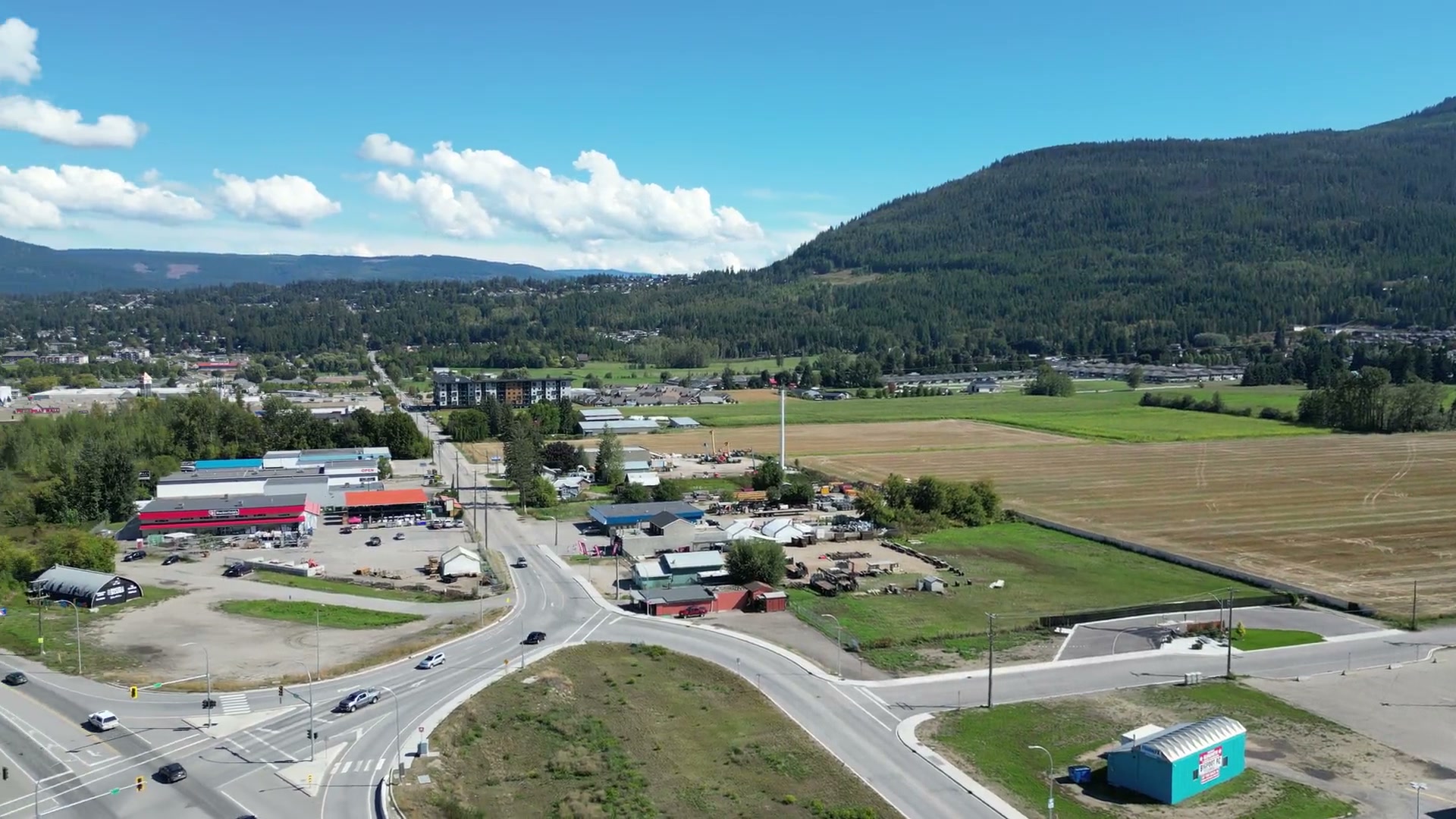 Turning from the roof of Westgate Public Market across the fields to a signalled intersection at the west side of Salmon Arm, and the highway running on into town under a forested hill