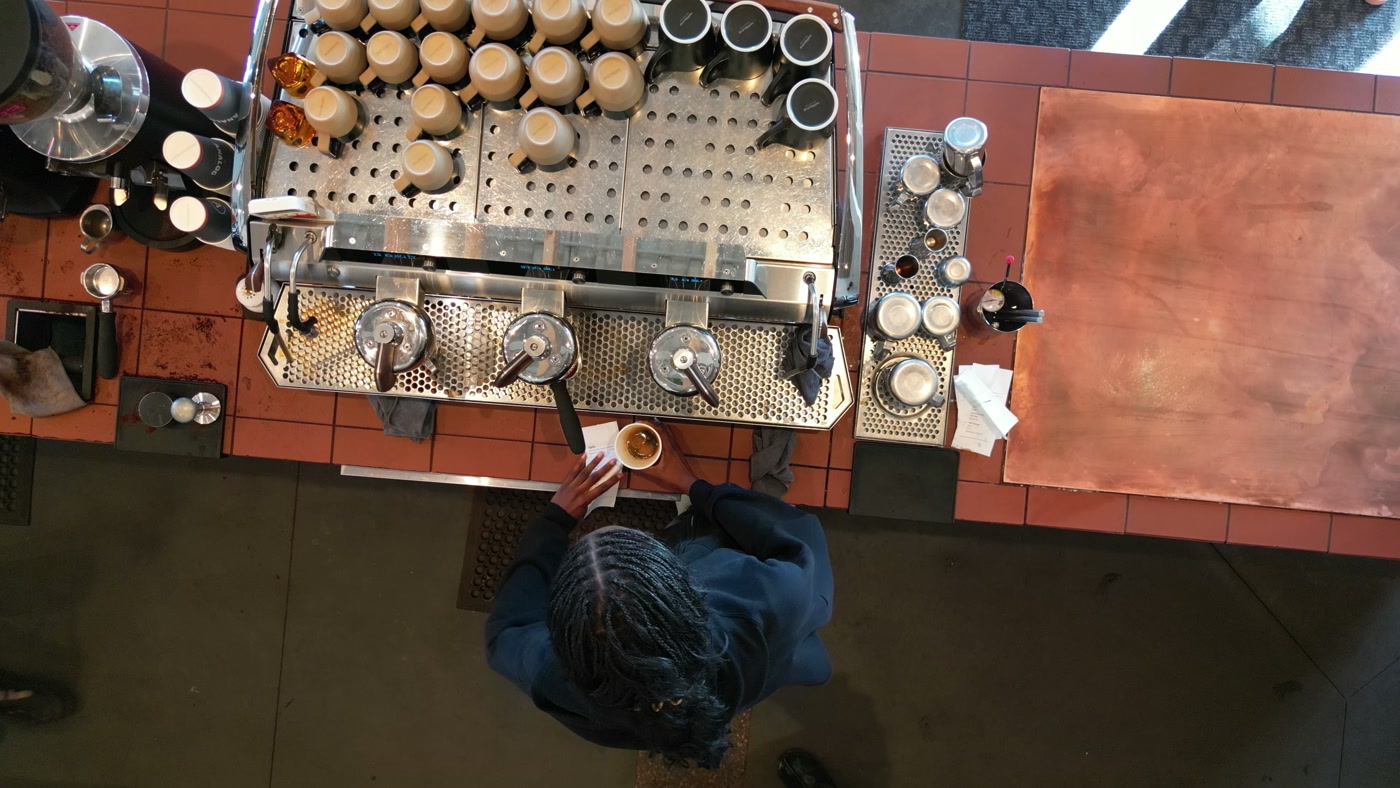 The espresso bar from directly overhead: the machine's perforated steel top stacked with cups, the copper-tiled counter, two baristas either side