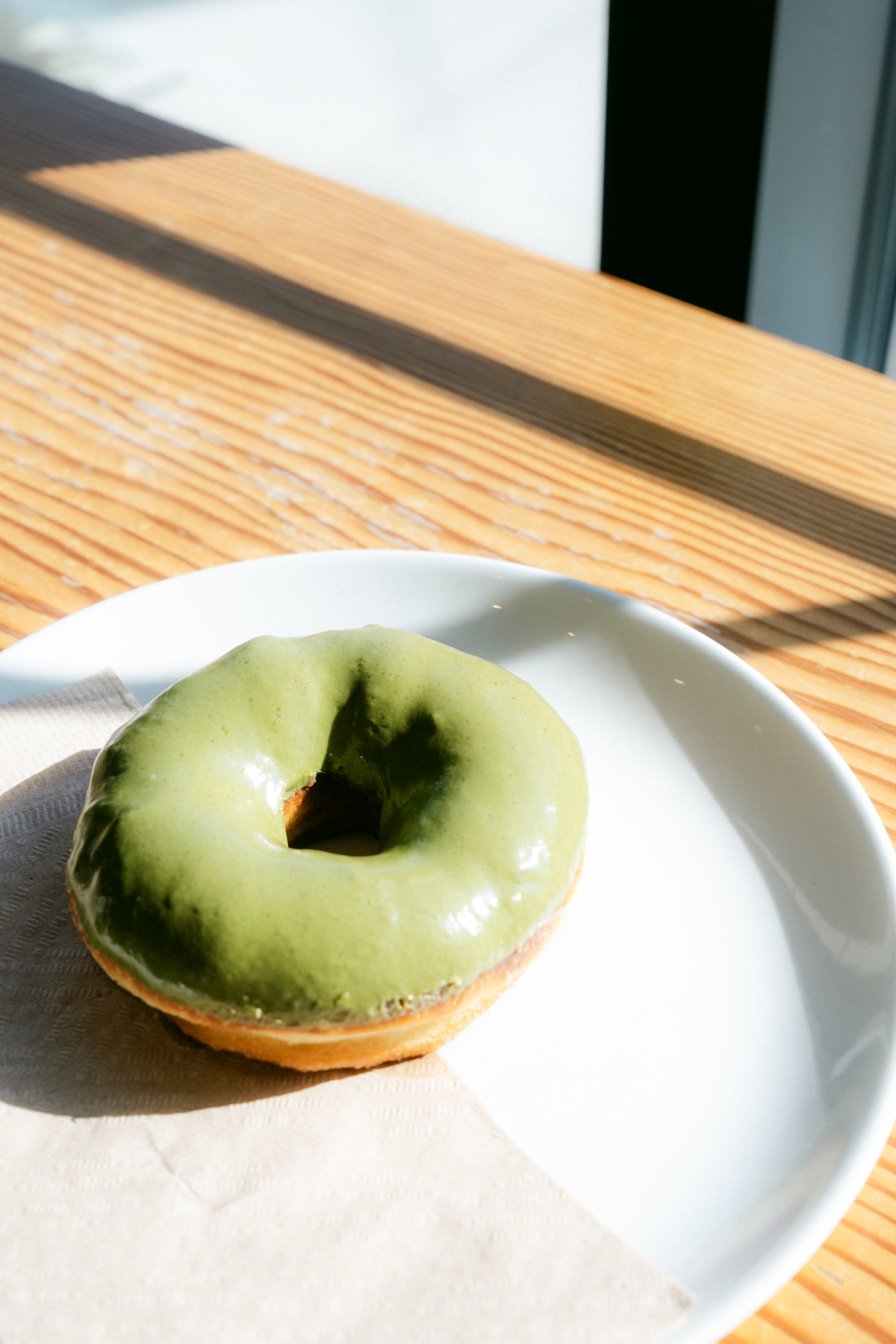A matcha-glazed doughnut on a white plate in hard morning sun on the cafe's patio table