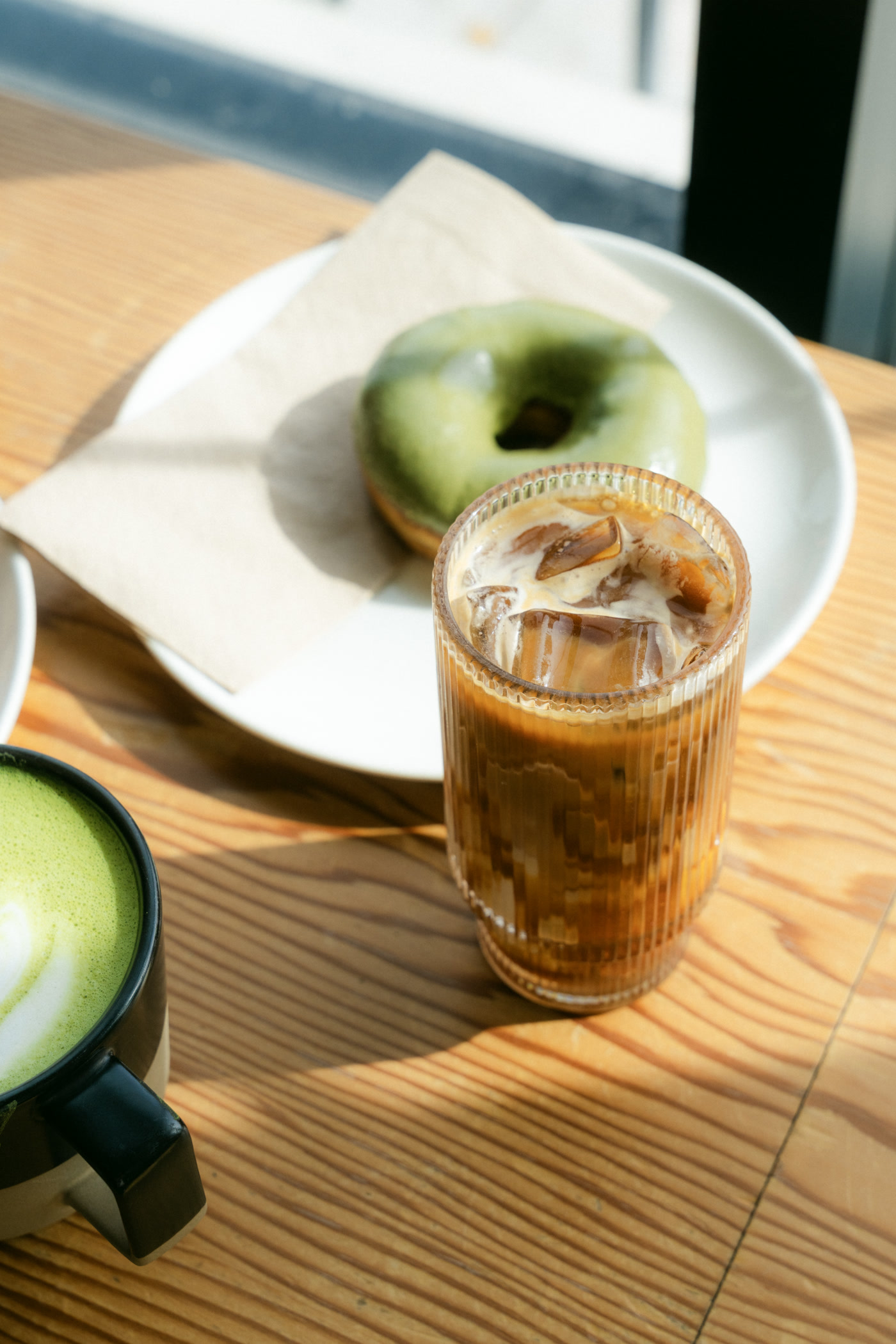 The matcha latte's poured leaf in a dark mug, the iced coffee and the doughnut behind it