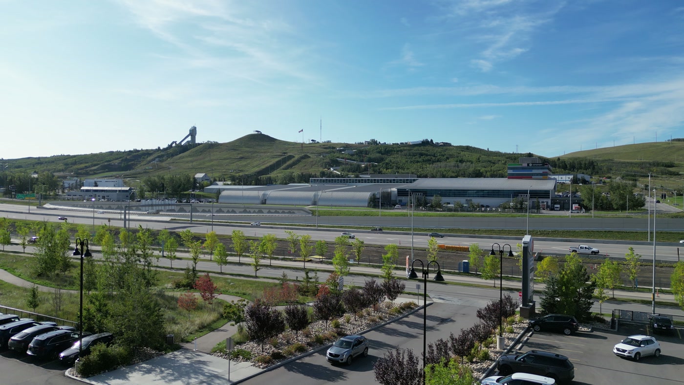 Canada Olympic Park across the highway from the cafe: the ski jump tower on the green shoulder, a bobsleigh track curving down through the grass, a flag at the summit and WinSport's arched roofs at the foot