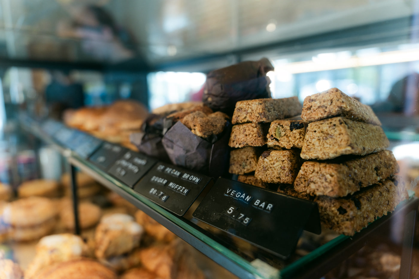 The pastry case from the side: scones and loaves on the glass shelf, price cards for a sugar muffin and a vegan bar