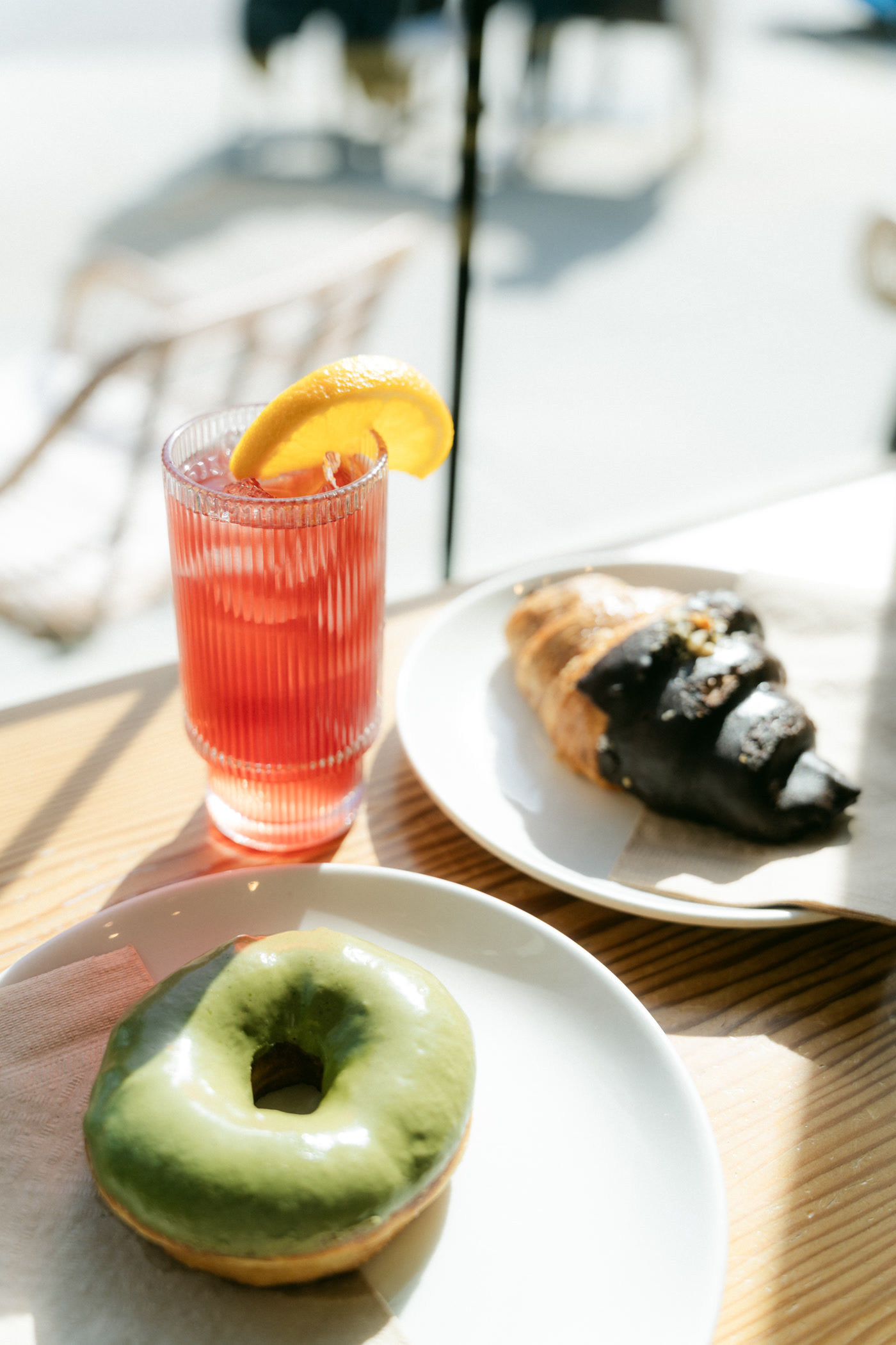The patio table in full sun: a red iced drink with an orange slice, the chocolate croissant and the matcha doughnut