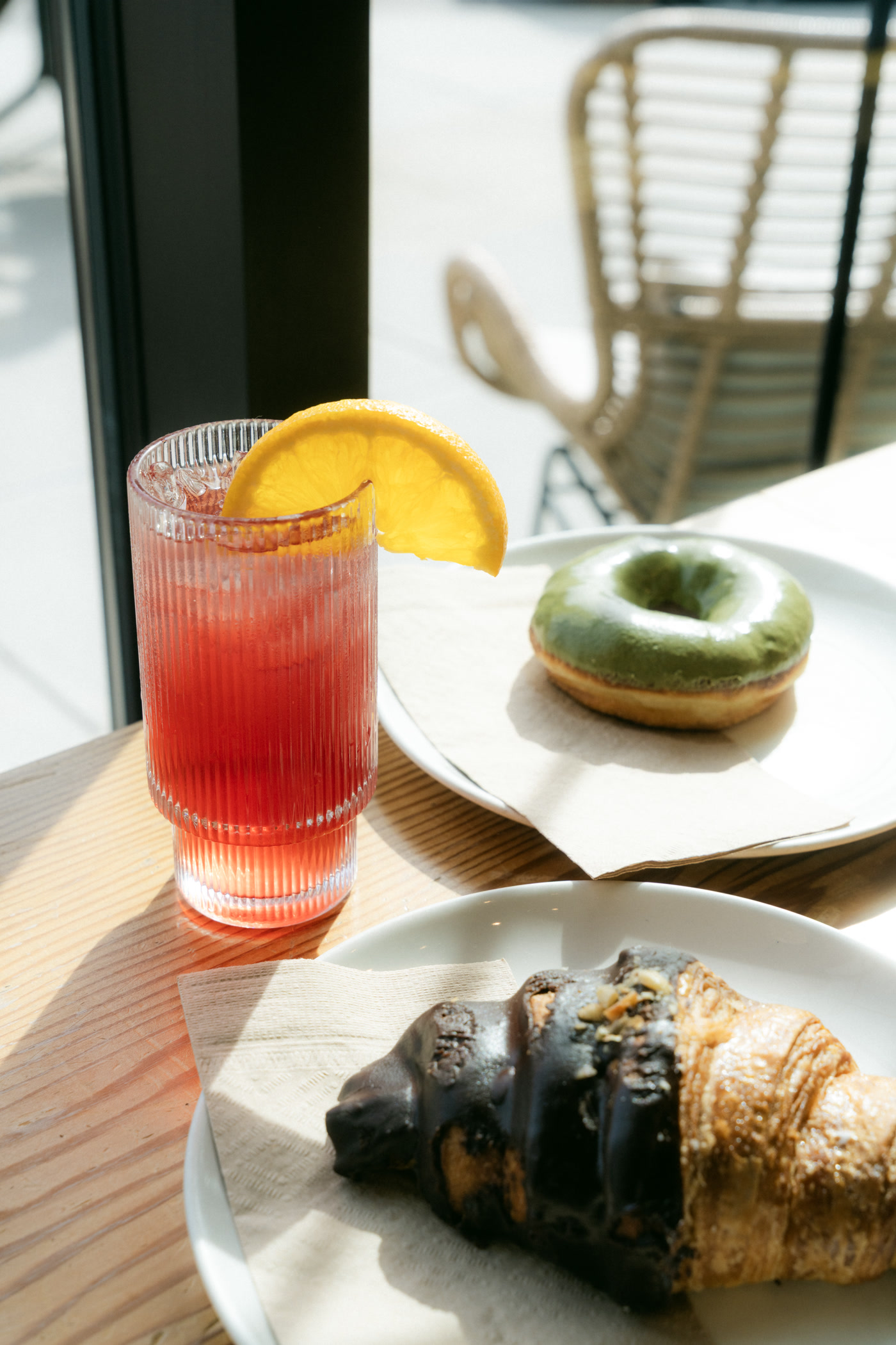 The patio table from above in hard sun: the red iced drink, the doughnut, the croissant and the latte, a patio chair behind