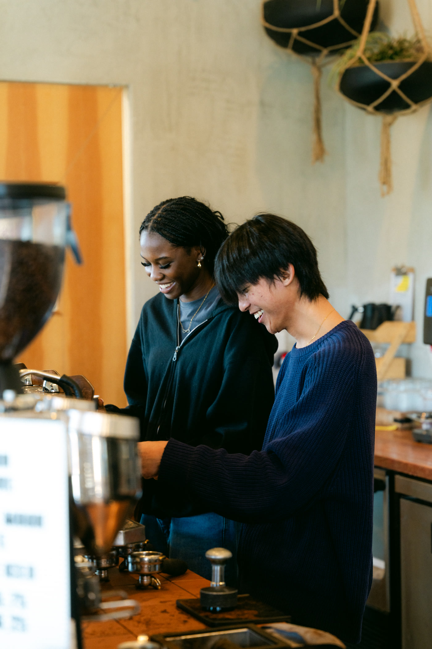 Two baristas working shoulder to shoulder behind the bar, the bean hopper in the foreground