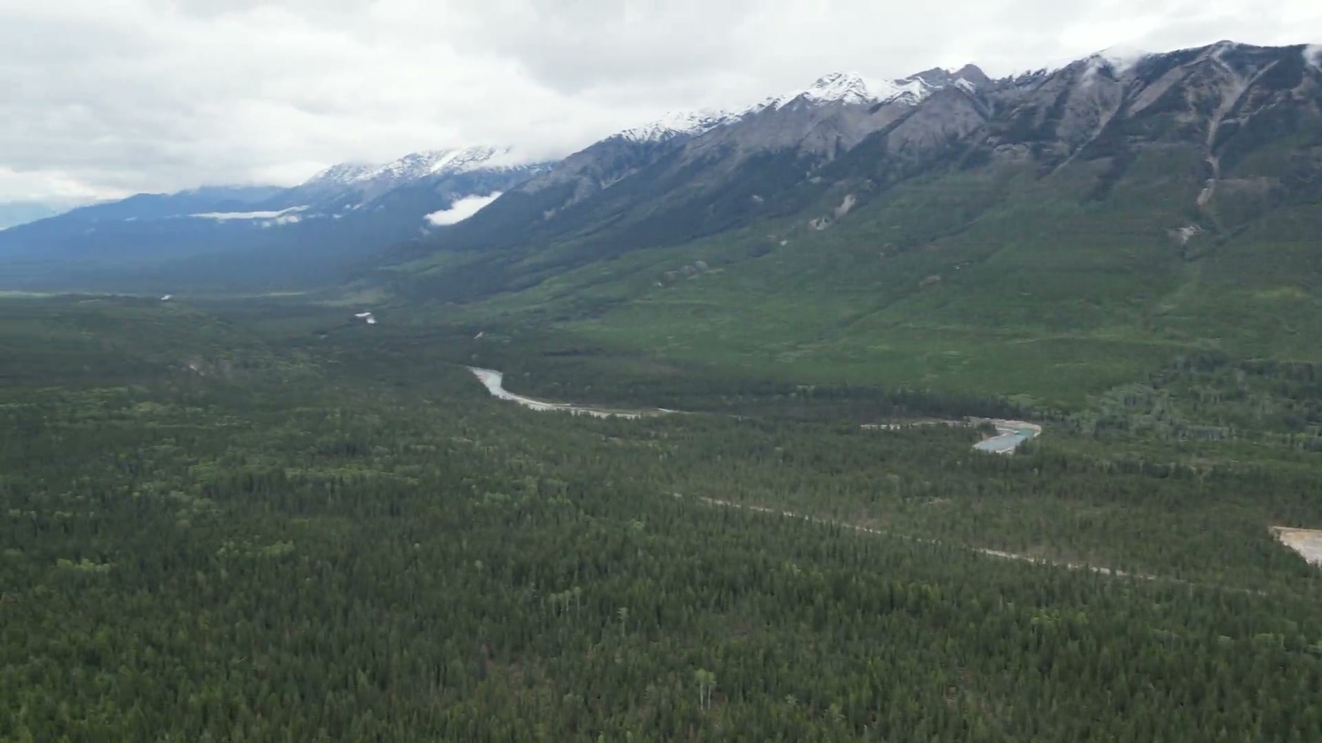 A snow-dusted summit ridge above the forested floor of the Columbia valley at Radium Hot Springs, scree chutes down its face, a gravel working and a turquoise pond among the trees below