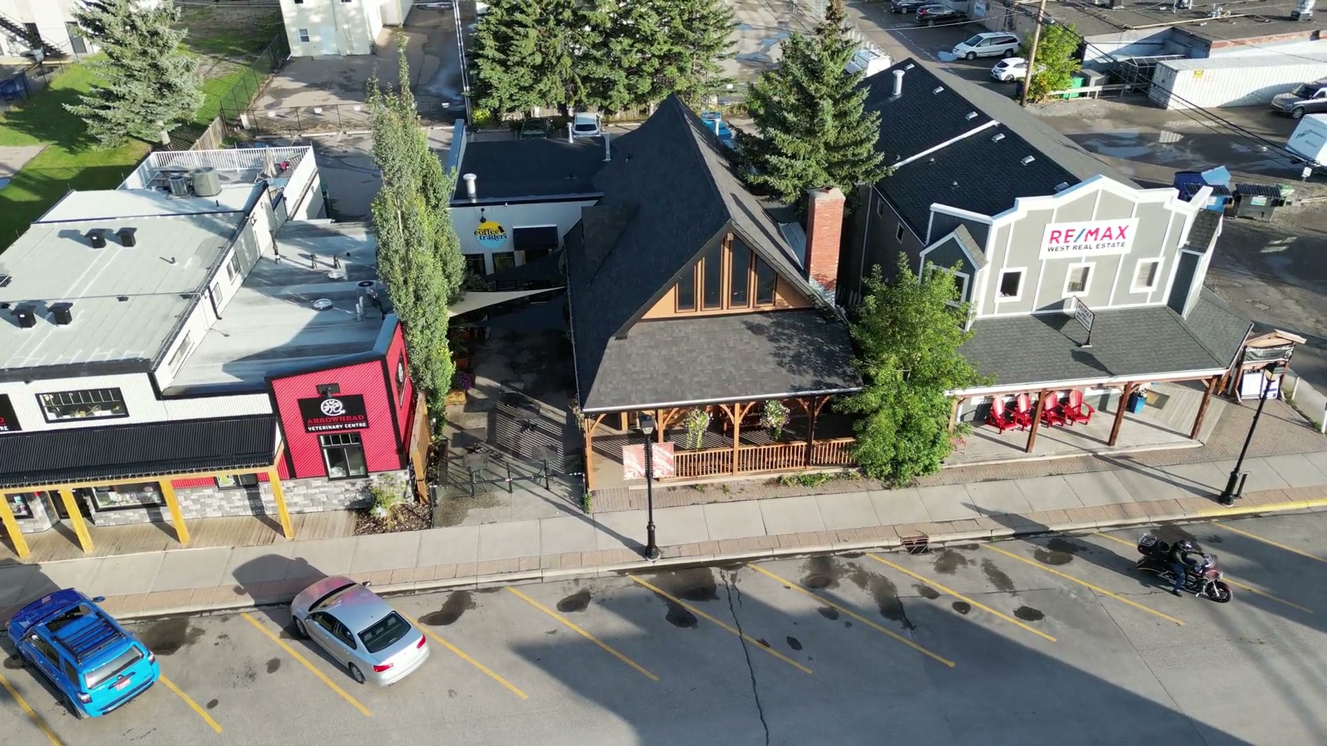 Descending at an angle toward Cochrane Coffee Traders on 2nd Avenue West: its dark gable roof and red-brick chimney, the courtyard of shade sails, parked cars, a motorcyclist and long shadows across the street