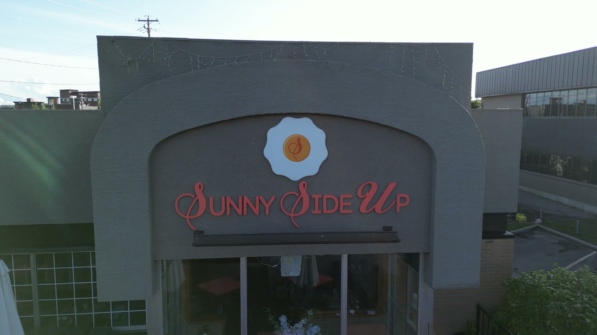 The camera at parapet height, hovering before the Sunny Side Up sign in Cochrane: the scalloped white egg, the script S in its yolk, the red letters beneath