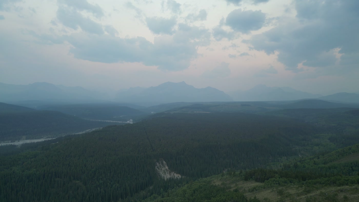 Blue hour from a summit in the Alberta foothills: forested ridges falling to a valley with a braided river, mountains on the western horizon and heavy cloud lit pink from beneath