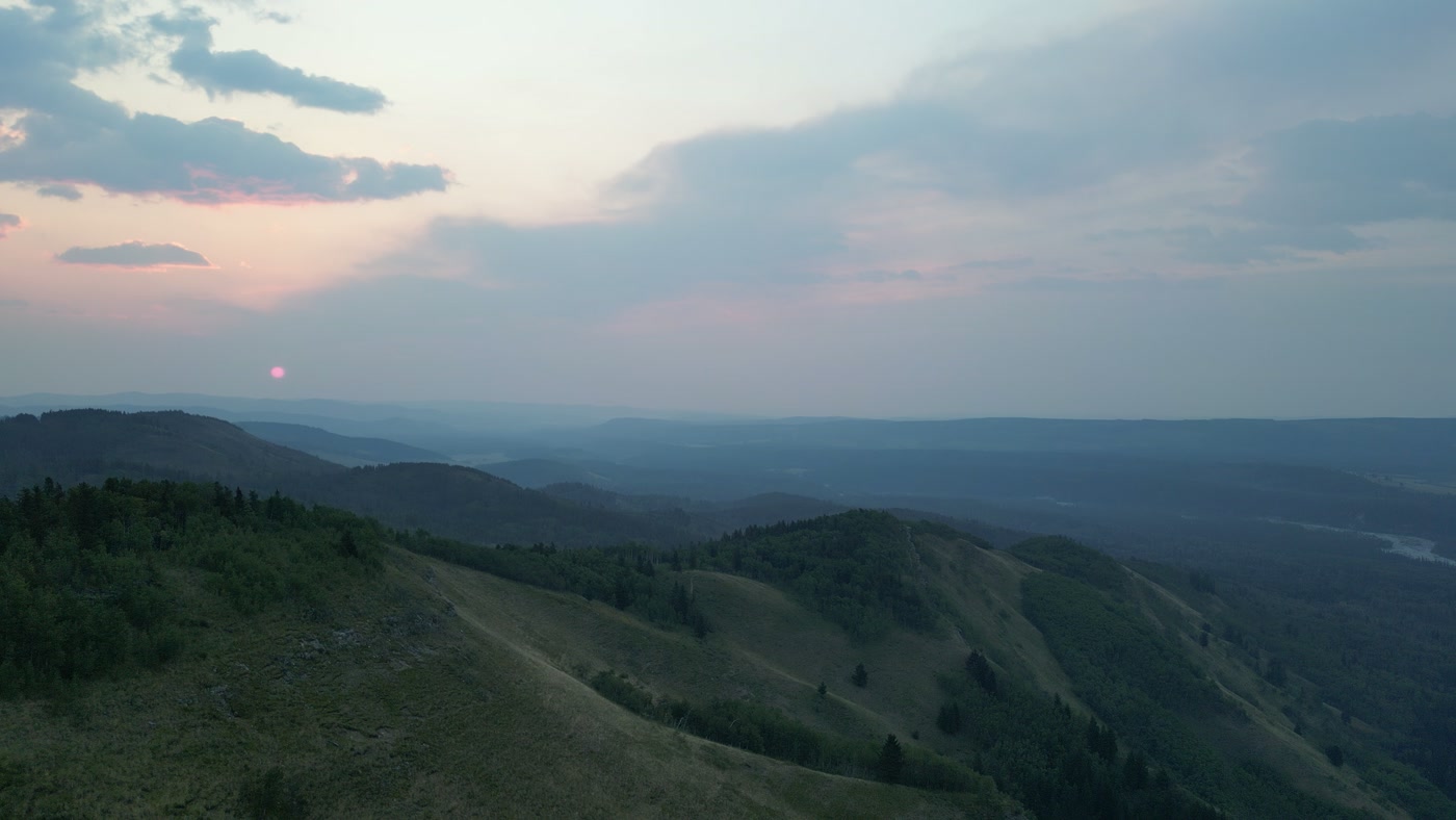The sun just clear of the last ridge at sunrise, a pink-red disc in the haze, the layered forested ridges of the Alberta foothills falling away below it