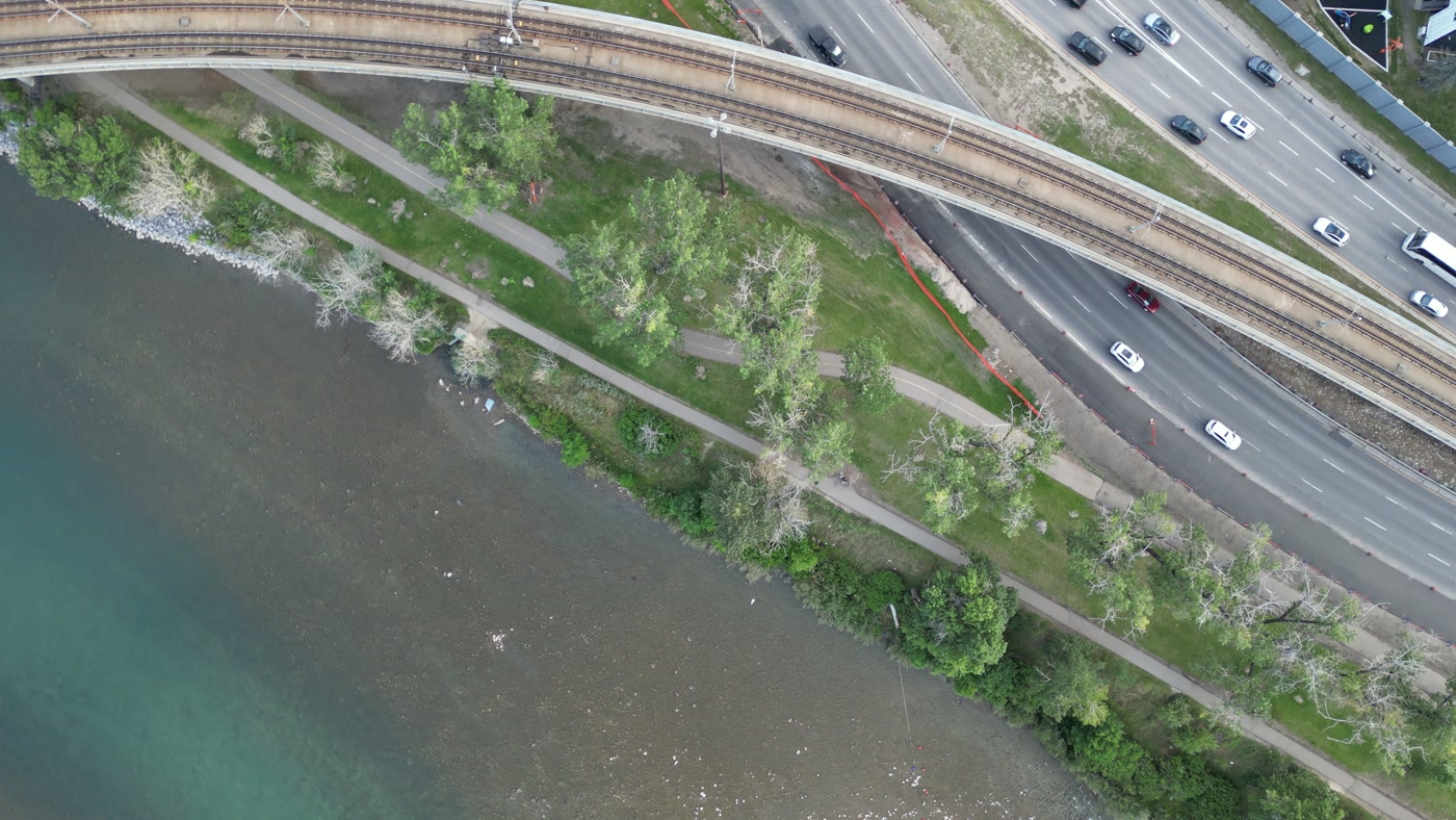 Memorial Drive from directly above: the CTrain's elevated track across the top of the frame, the lanes, the tree-lined pathway and the stony edge of the Bow River