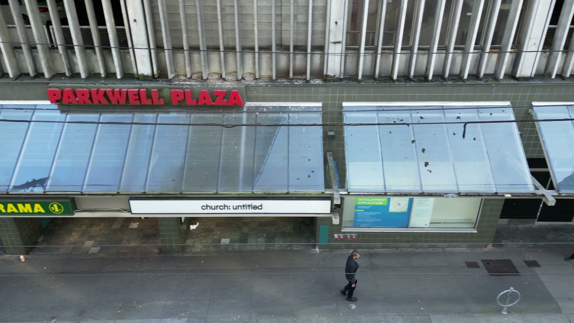 Sideways along the glass canopies of a storefront row on Seymour Street, past a green neon sign and a PhoneBox, to PARKWELL PLAZA in red letters, DOLLARAMA and the church: untitled fascia