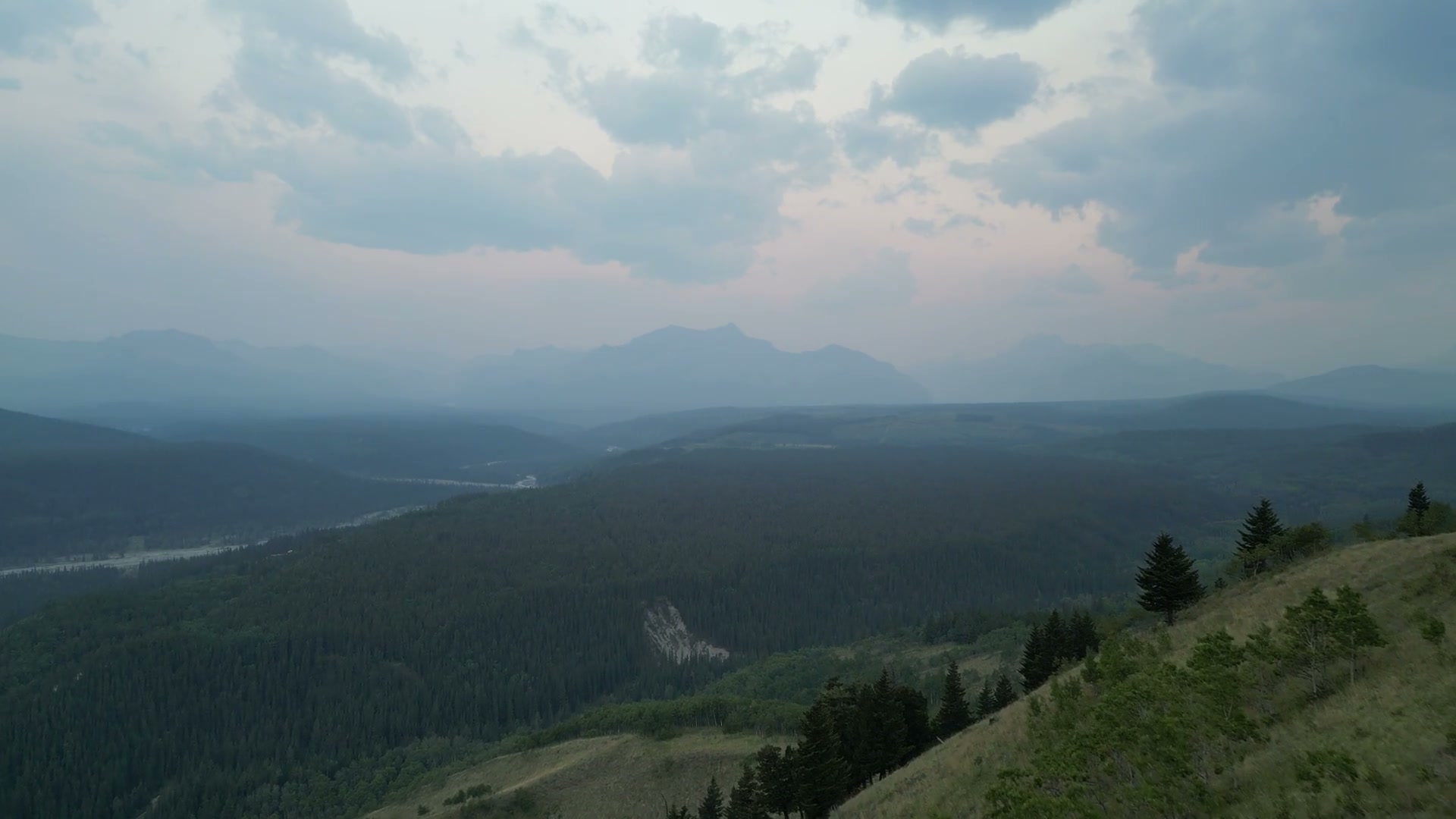 Pulling back from a grass crest with two small pines to a blue-hour valley in the Alberta foothills: a braided river, forested ridges and mountains under cloud lit pink from beneath