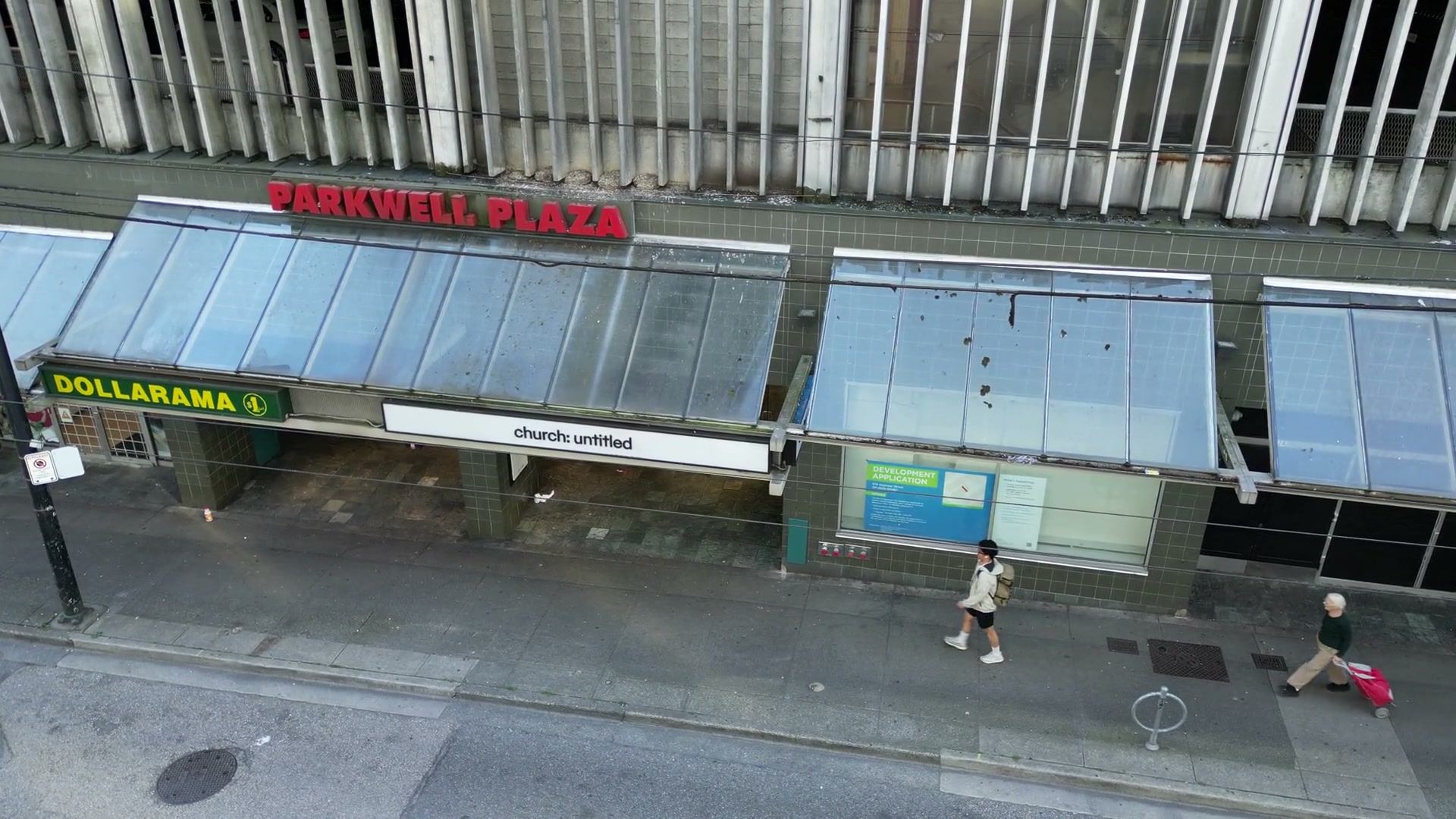 A quick pan right to left along the glass canopies on Seymour Street, settling on PARKWELL PLAZA, a DOLLARAMA sign and the white fascia of church: untitled while people pass on the sidewalk