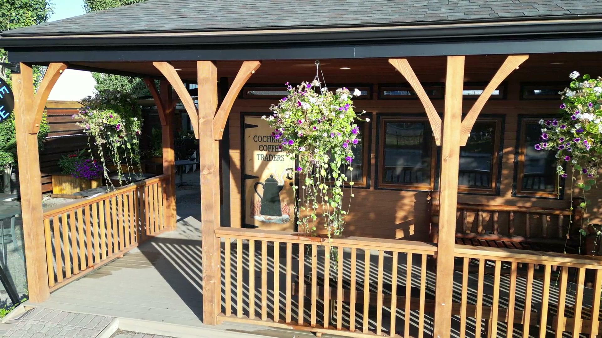 Sliding slowly left along the timber porch of Cochrane Coffee Traders on 2nd Avenue West as its painted name clears a hanging basket and a round black Coffee House sign enters the frame