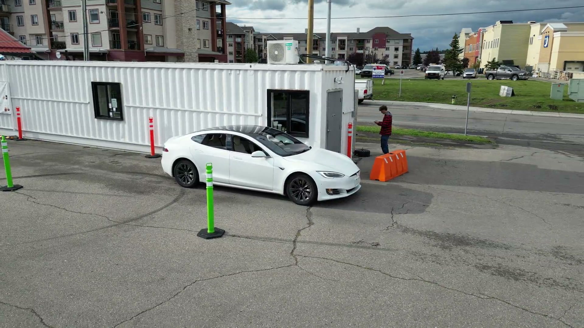 The camera flying low beside the drive-thru lane of Covet Sips + Sweets in Cochrane, following a white car past the menu banner on the white shipping container to the first window, then on to the window at the end, where someone leans out toward it