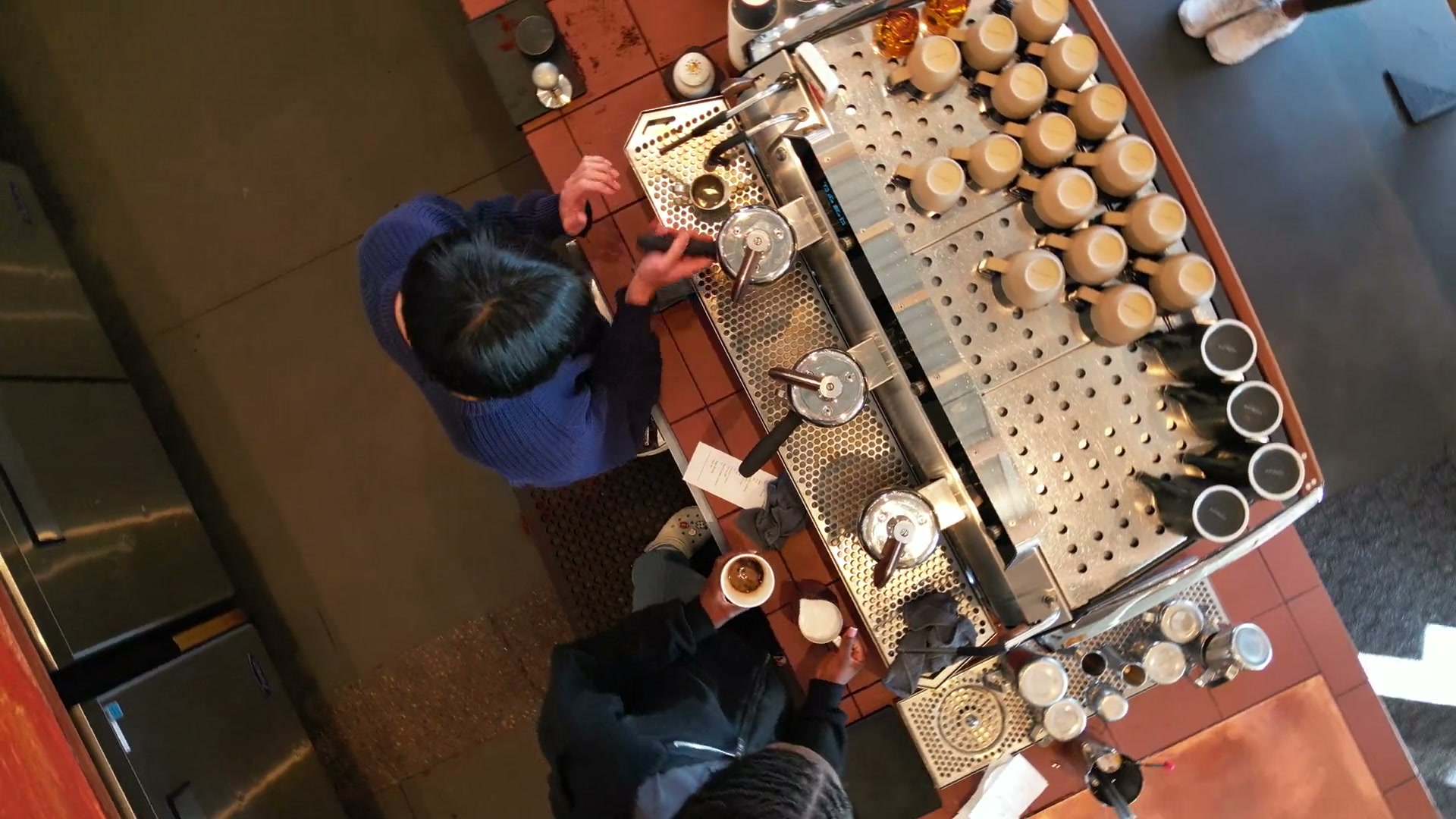 Directly above a café's espresso bar, turning slowly: two baristas at the machine, rows of cups along the counter