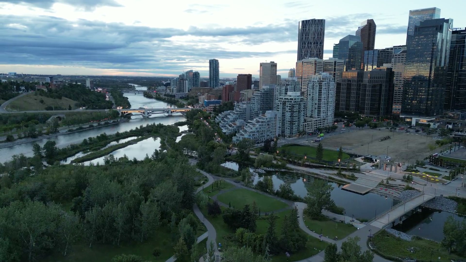 Slow pull-back over the Bow River and Prince's Island toward downtown Calgary at dusk, towers catching the last light under a broken cloud