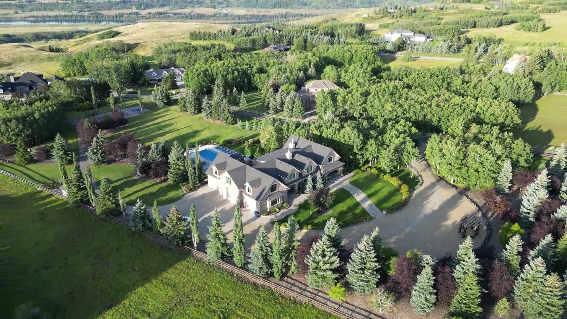 Slow pull-back from the roofline of a large estate home with a pool, revealing treed grounds and open Alberta country beyond