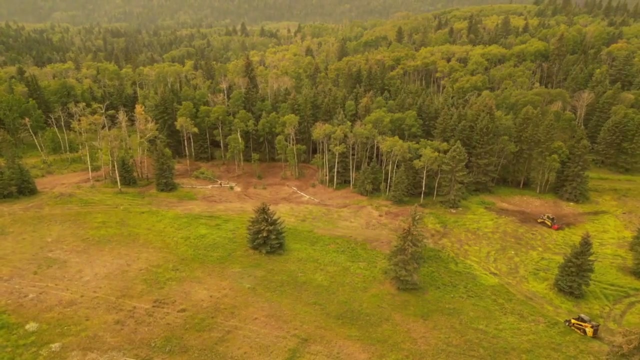 Aerial pass over a foothills acreage in late summer, machinery working the edge of the trees