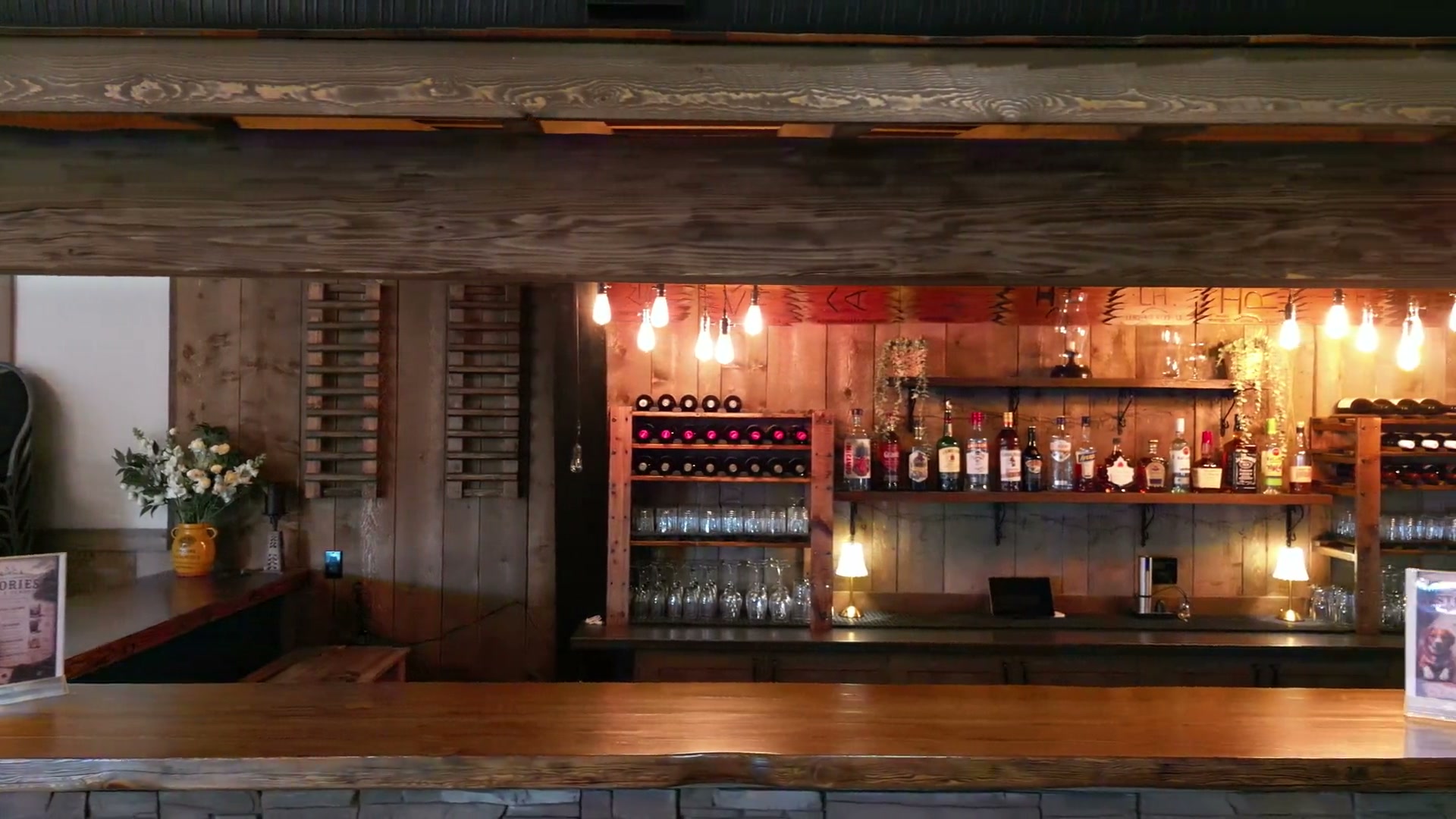 The venue's bar under heavy timber beams: bottles, glassware and warm bulbs