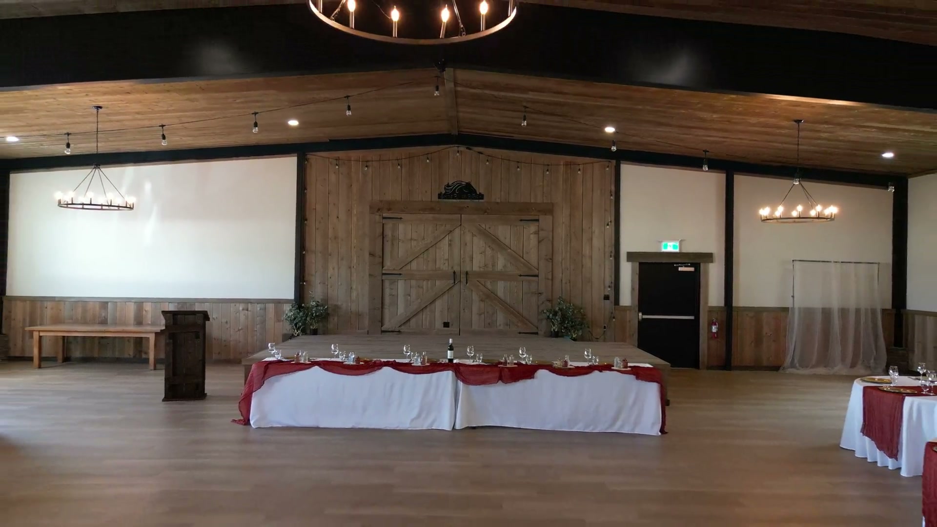 Pulling back from the barn doors of a timber wedding hall: the head table, a chandelier, round tables set in white and burgundy